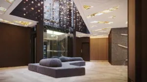 Moderni viešbučio fojė su pilka sofa, stikliniu liftu ir kabančiomis šviečiančiomis dekoracijomis lubose.