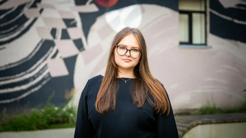 Jauna ilgaplaukė moteris su akiniais vilkinti juodą palaidinę stovi fone su abstrakčiu gatvės meno piešiniu.