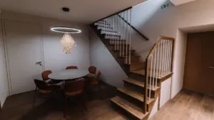 Moderni valgomasojo zona su apvaliu stalu, kėdėmis bei mediniais laiptais šviesiame interjere.