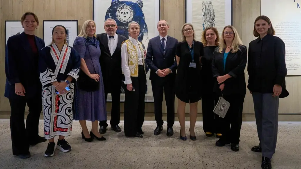 Grupė elegantiškai apsirengusių žmonių stovi galerijoje priešais meno kūrinius su meškos atvaizdu.