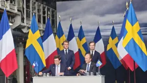 Prancūzijos ir Švedijos lyderiai pasirašo susitarimus prie stalo su abiejų šalių vėliavų fonu.