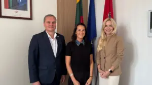 Trys besišypsantys žmonės stovi kabinete priešais Lietuvos, Europos Sąjungos ir rajono vėliavas.