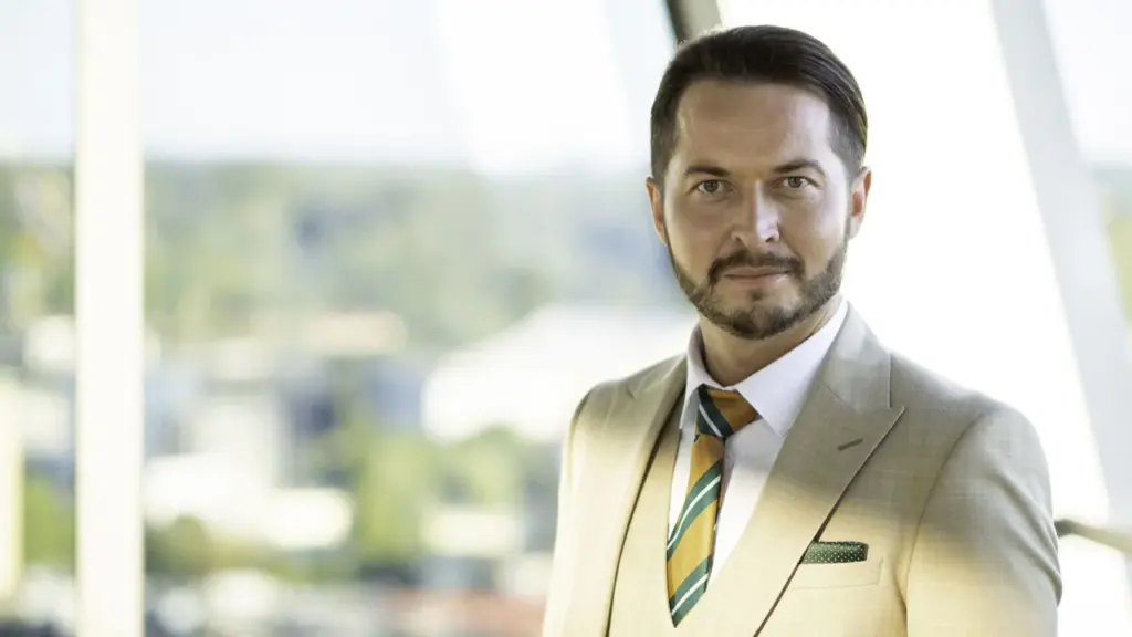 Vidutinio amžiaus barzdotas vyras šviesiu kostiumu stovi biure prie didelio stiklinio lango.