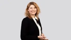 Besišypsanti šviesiaplaukė moteris vilkinti baltus marškinius ir juodą švarką pilkame fone.
