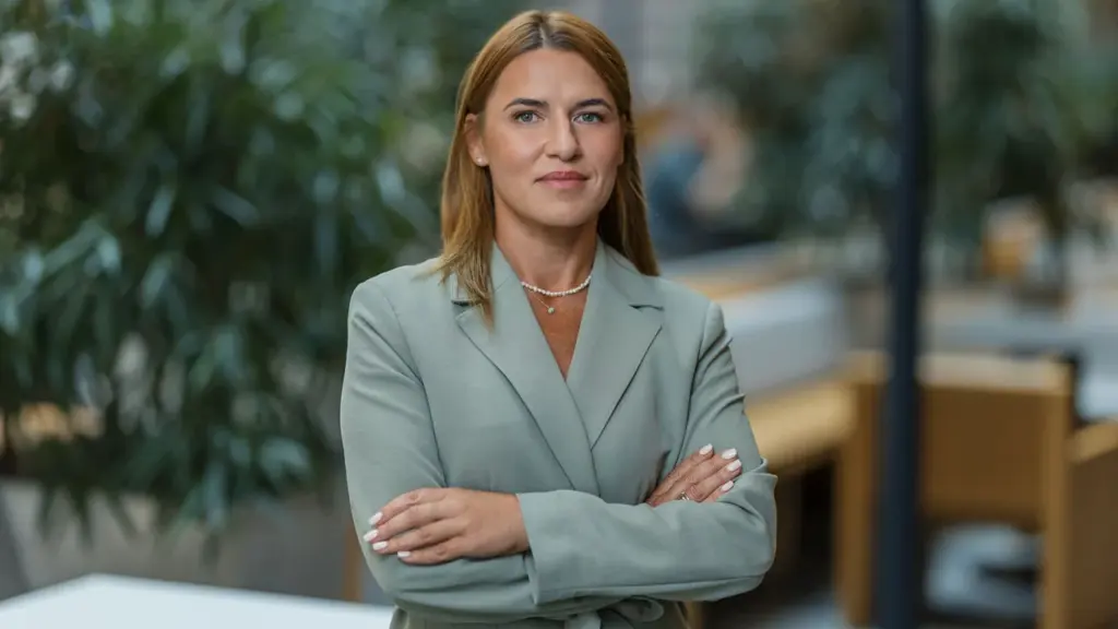Šviesiaplaukė moteris šviesiai žaliu švarku stovi biure sukryžiuotomis rankomis ir žvelgia į kamerą.